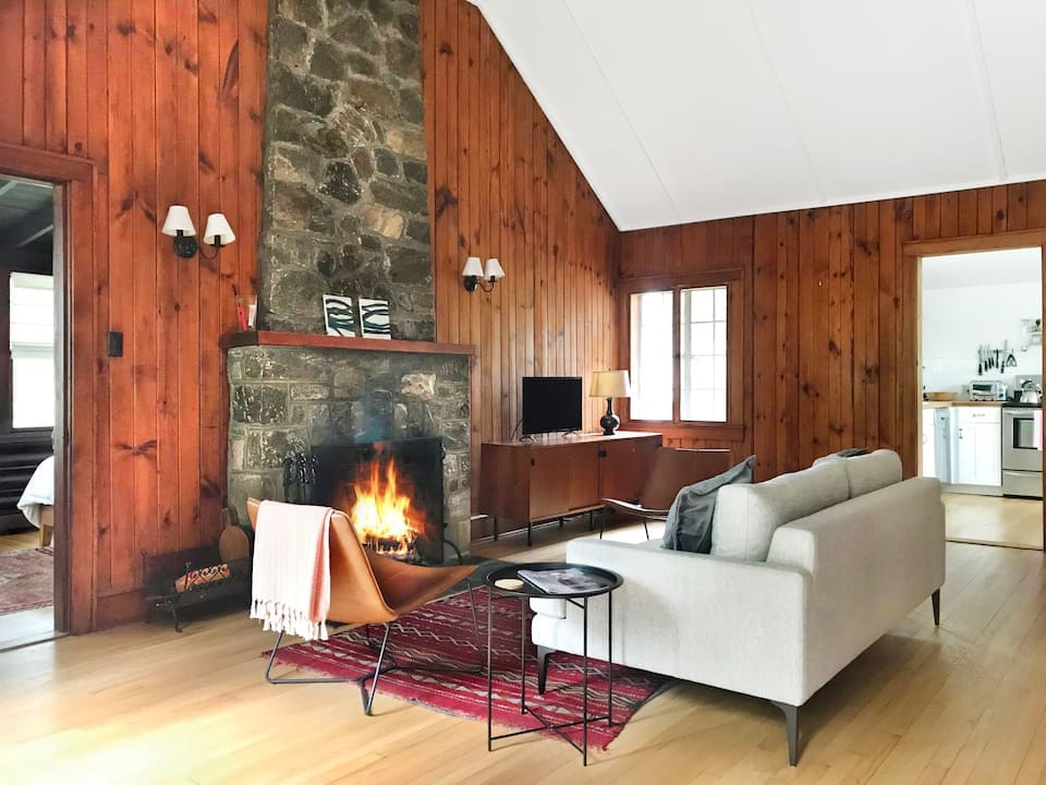 A cozy living area is highlighted by a stone fireplace with a lit fire, surrounded by wooden walls. A light-colored sofa faces the fireplace, while a small side table and a textured rug add warmth to the space. Nearby, a kitchen is partially visible.