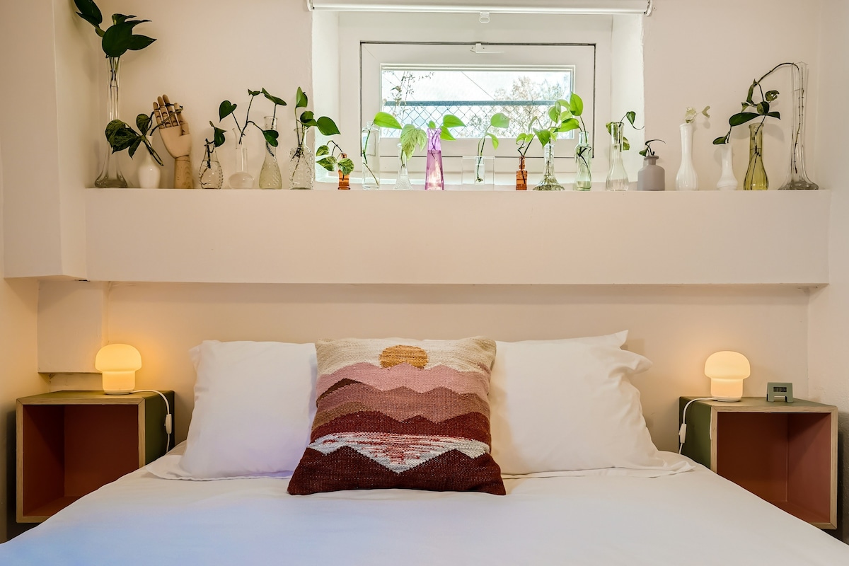 A cozy bedroom is highlighted by a spacious bed, adorned with white linens and a decorative pillow featuring mountain motifs. Soft lighting from two bedside lamps creates a warm ambiance, while a collection of green plants in various vessels sits on the shelf above the bed.