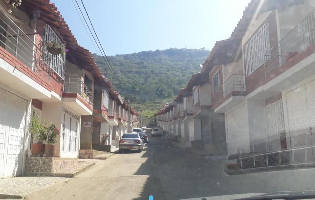 A view of a serene neighborhood lined with two-story houses features red-tiled roofs and balconies. A narrow roadway runs through the center, with parked vehicles on either side. Lush green hills rise in the background under a clear blue sky.