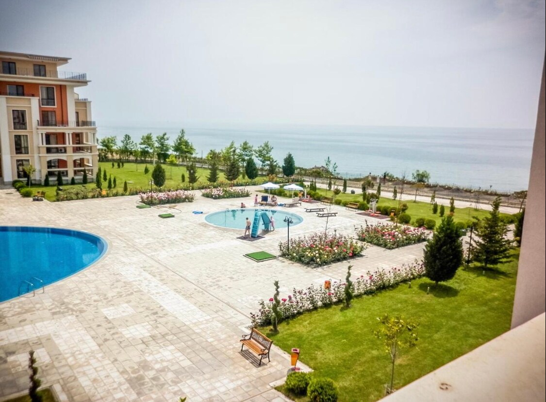 A spacious outdoor area features a large swimming pool surrounded by landscaped gardens and vibrant flower beds. The sea can be seen in the background, enhancing the serene setting. Several lounge chairs are arranged around the pool area, inviting relaxation.