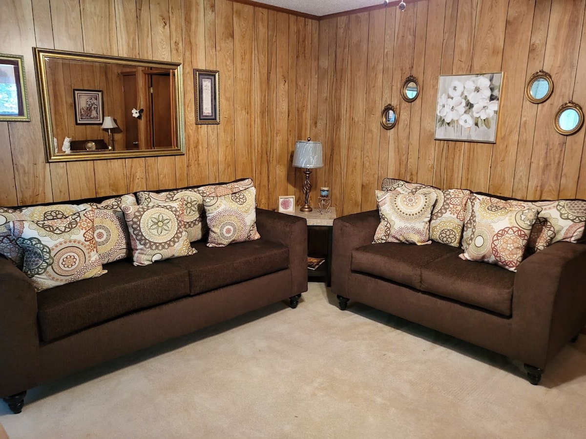 Two brown sofas with decorative pillows sit opposite each other, framed by wooden paneling. A small table is positioned between them, highlighted by a lamp. A mirror and floral artwork adorn the wall, reflecting a cozy and inviting space.