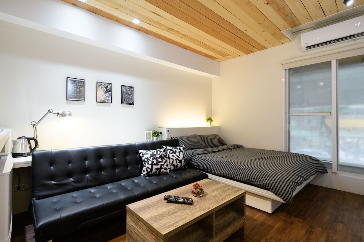 A contemporary studio space features a spacious bed with a striped duvet and a stylish black sofa. A wooden coffee table occupies the center, complemented by small decorative plants. Soft lighting from above highlights the warm wood-paneled ceiling, while a large window allows natural light to enter.