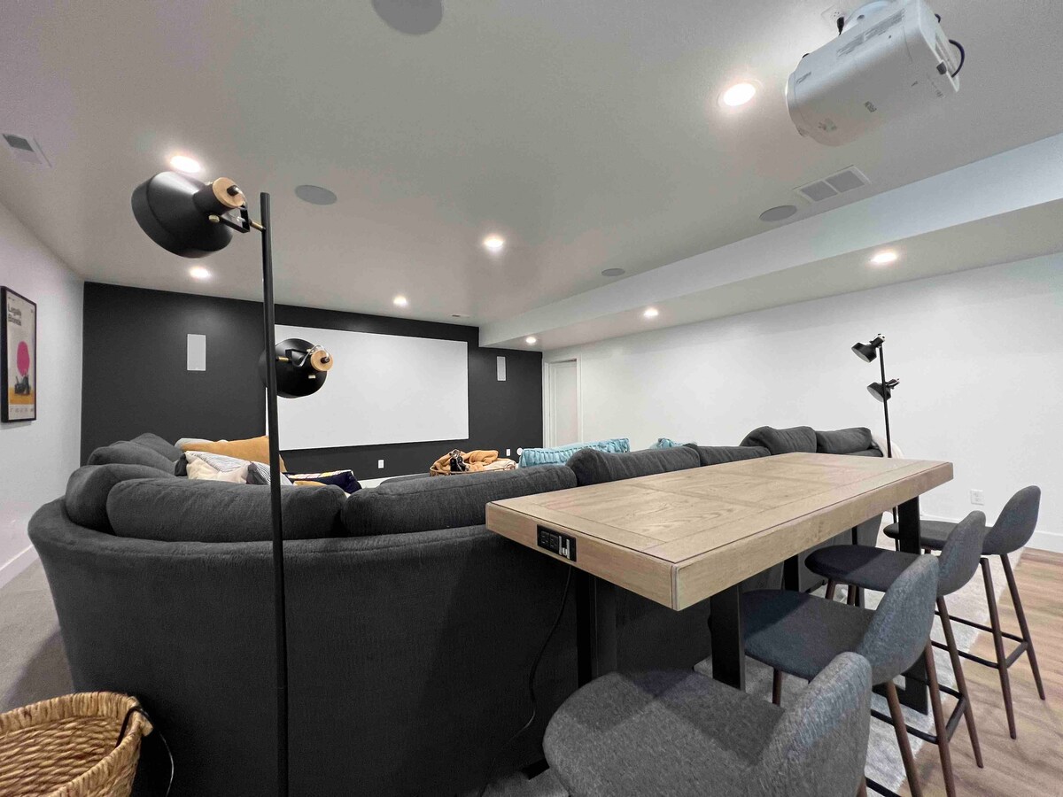A spacious theater room features a large, curved sectional sofa arranged around a central coffee table. The wall is equipped with a projector screen, while built-in speakers and overhead lights enhance the viewing experience. Additional seating is provided by modern chairs at a nearby table.