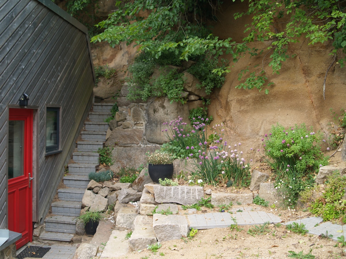 A well-maintained entrance features red double doors next to stone steps leading to an outdoor area. Lush greenery is complemented by colorful flowering plants in black pots along the rocky wall, creating a peaceful and inviting approach to the home.
