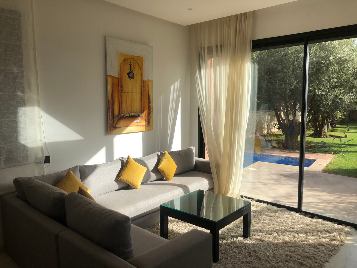 A light-filled living area features a spacious grey sectional sofa adorned with yellow cushions. A glass coffee table is positioned in front of the seating. Sheer curtains softly filter sunlight, while a view of the garden and private pool is framed by large sliding glass doors.
