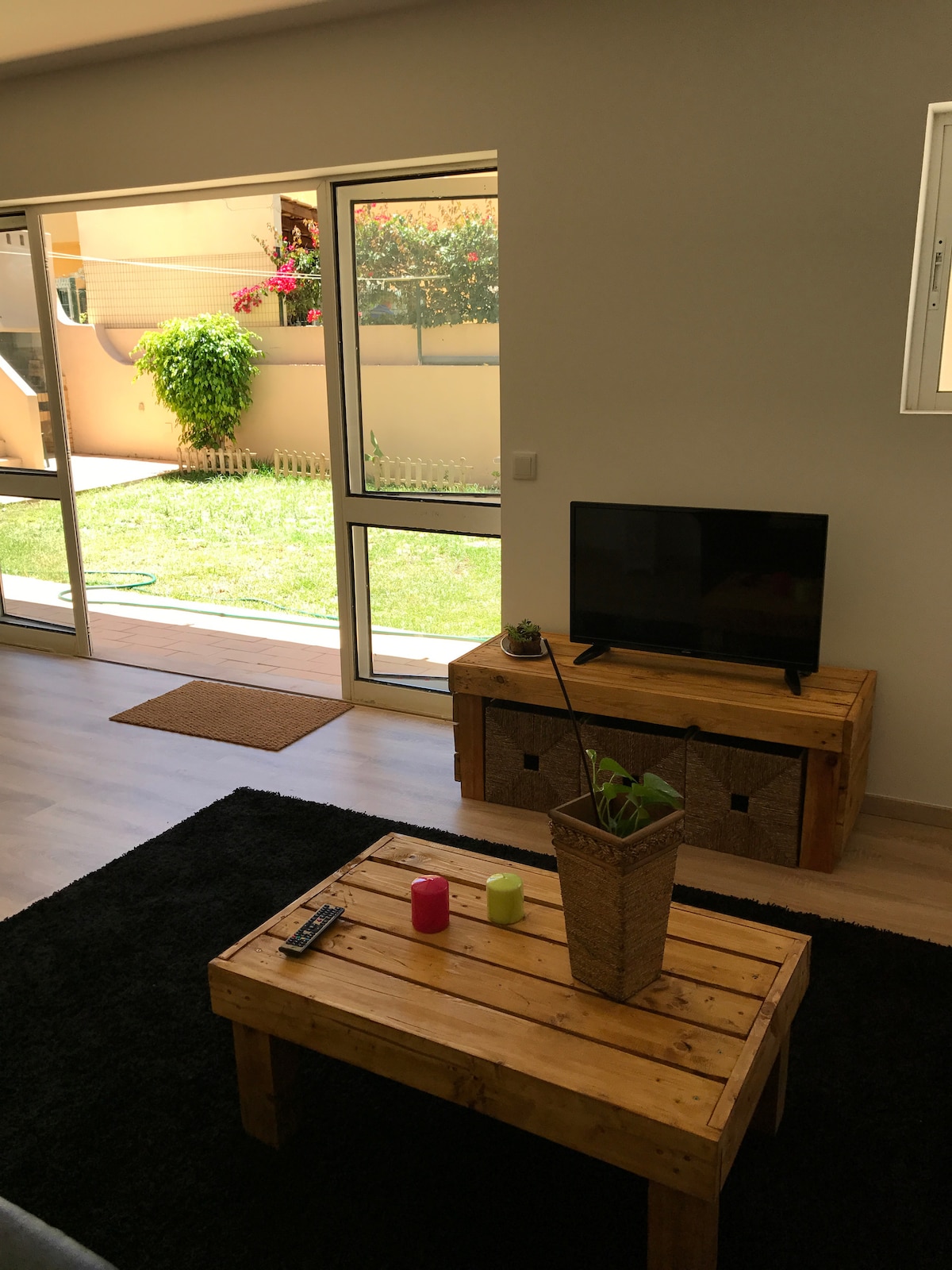 A living area is depicted, featuring a wooden coffee table and a flat-screen television on a wooden stand. Large glass doors open to a green outdoor space, enhancing the light-filled atmosphere. Decorative items include a potted plant and candles on the table.