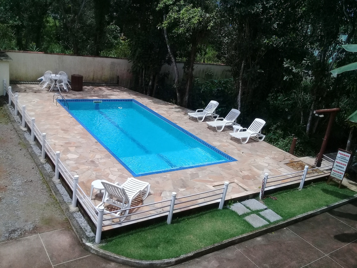 A rectangular pool is surrounded by stone tiles and tropical greenery. Several white lounge chairs are positioned along the pool's edge. A small grassy area features stepping stones leading to the pool, while a shaded seating area is visible nearby.