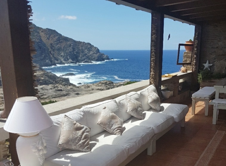 An inviting outdoor seating area features a white sofa adorned with decorative pillows, overlooking a rocky coastline. The blue sea and distant hills are visible in the background, while natural stone walls provide a rustic charm. Sunlight streams through the open space.