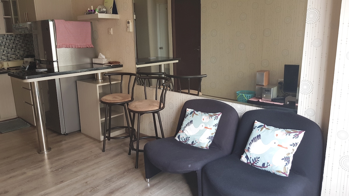 An open living area showcases a modern kitchen with stainless steel appliances, including a refrigerator. Two bar stools are positioned at a breakfast bar, while a dark sofa features two decorative cushions. Natural light streams in through sheer curtains, illuminating the space.