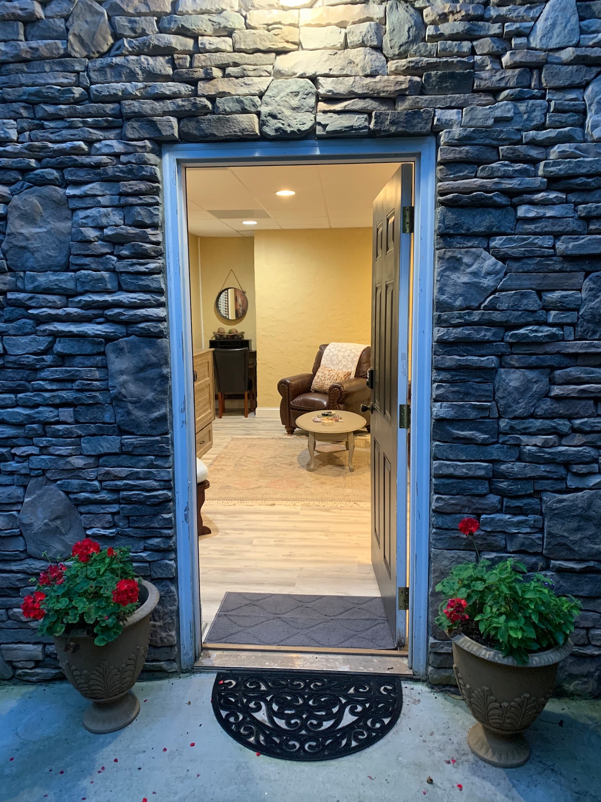 The entrance features a warmly lit doorway framed by textured stone walls. Potted red geraniums add a splash of color beside the entry. A comfortable seating area is visible through the open door, inviting guests into the suite.