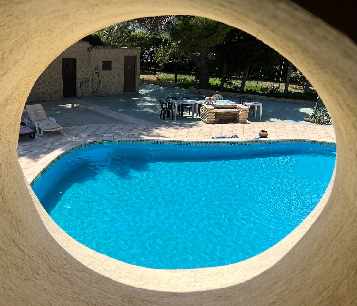 A view of the heated swimming pool is framed by an architectural opening. The pool's clear blue water reflects sunlight, and loungers are positioned nearby. A dining area is visible, with tables and chairs set under the shade of trees in the background.