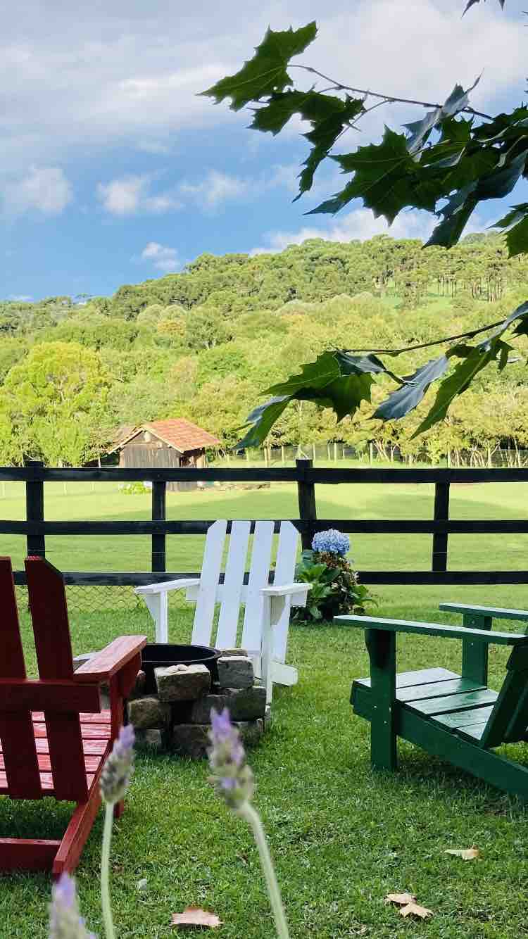 A spacious outdoor area features colorful adirondack chairs in red, white, and green, arranged around a stone fire pit. Lush green grass extends towards a scenic view of a rustic wooden barn and rolling hills, framed by trees under a partly cloudy sky.