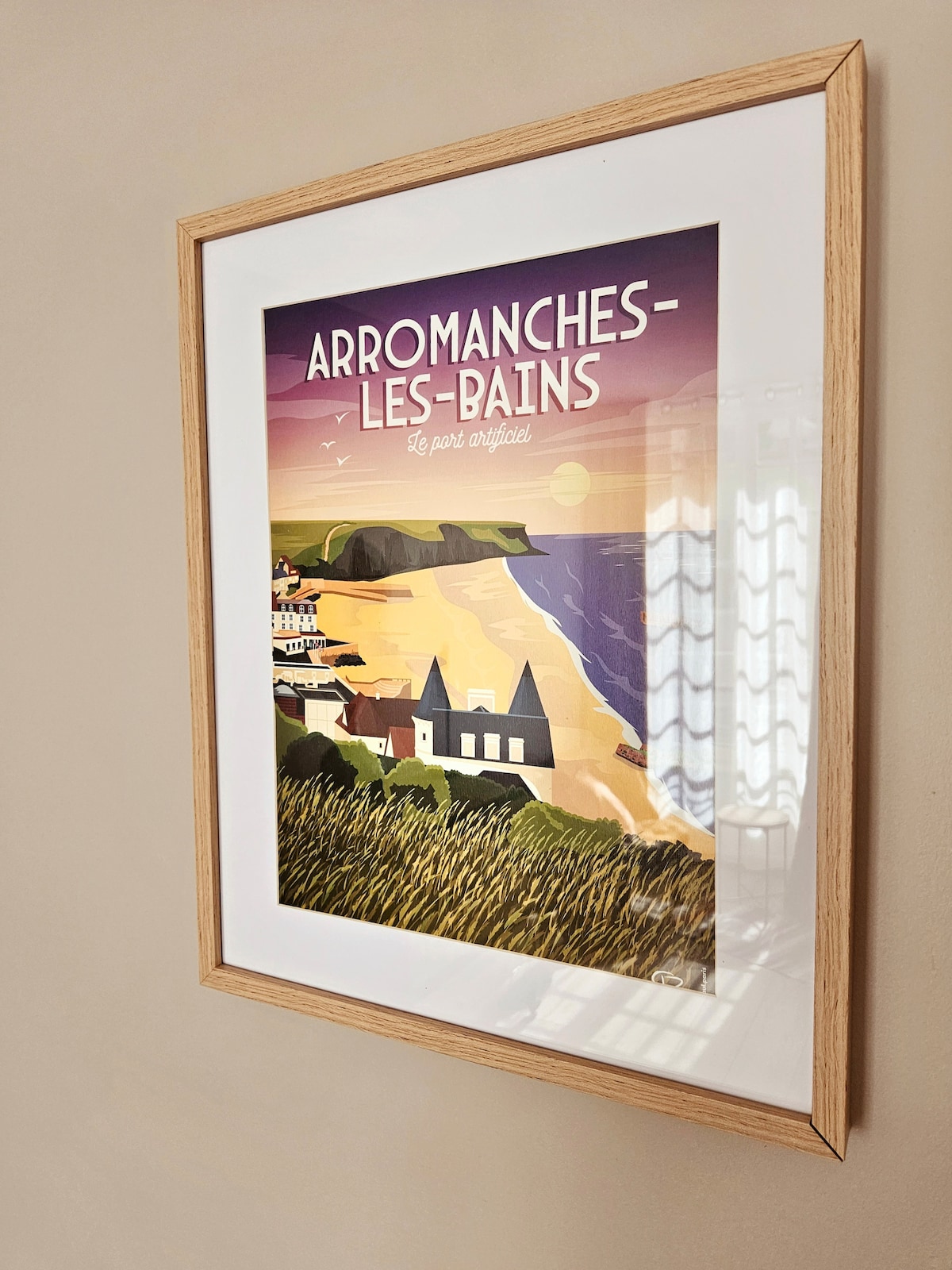 A framed vintage poster depicting Arromanches-les-Bains is hung on the wall. The artwork features a serene coastal landscape with soft pastel colors, showcasing a beach, cliffs, and a tranquil ocean under a sunset. A wooden frame surrounds the poster, adding a subtle touch to the decor.
