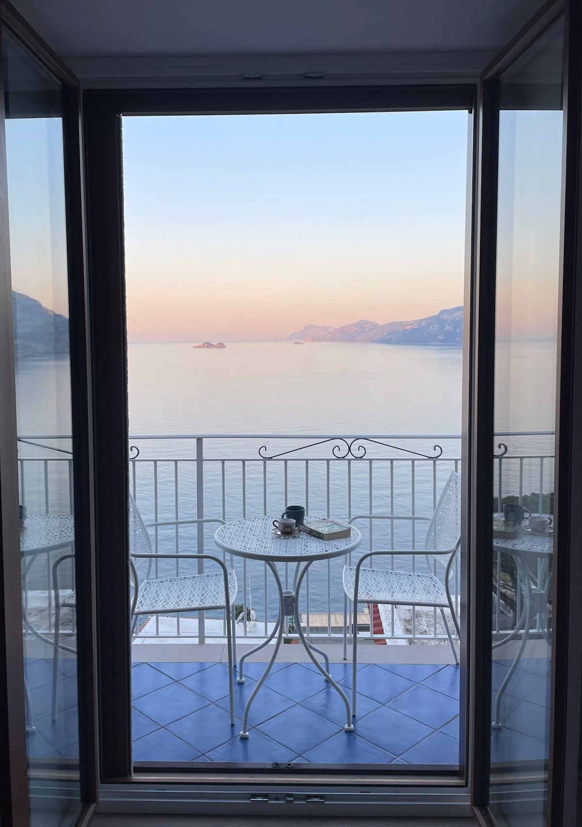 A serene balcony is framed by glass doors, offering a view of the tranquil sea at sunset. A small round table and two white chairs are positioned on the tiled floor, with soft pastel colors illuminating the horizon.
