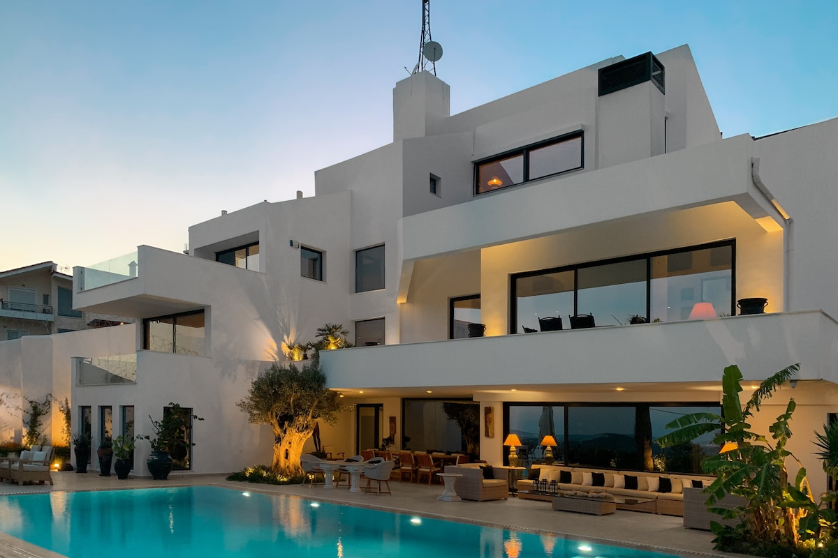 A contemporary villa is presented at twilight, showcasing its luminous white exterior and expansive windows. The swimming pool reflects the soft light of the evening, while outdoor seating areas invite relaxation. Lush greenery and decorative planters accentuate the serene atmosphere surrounding the space.