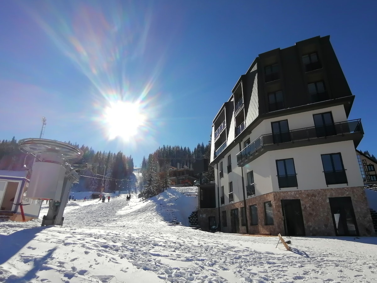 The exterior of a modern aparthotel is seen against a clear blue sky. Sunlight reflects off the snow-covered slope, revealing the nearby ski lift and tree-lined hillside. The building features a mix of dark and light materials, with balconies overlooking the winter landscape.