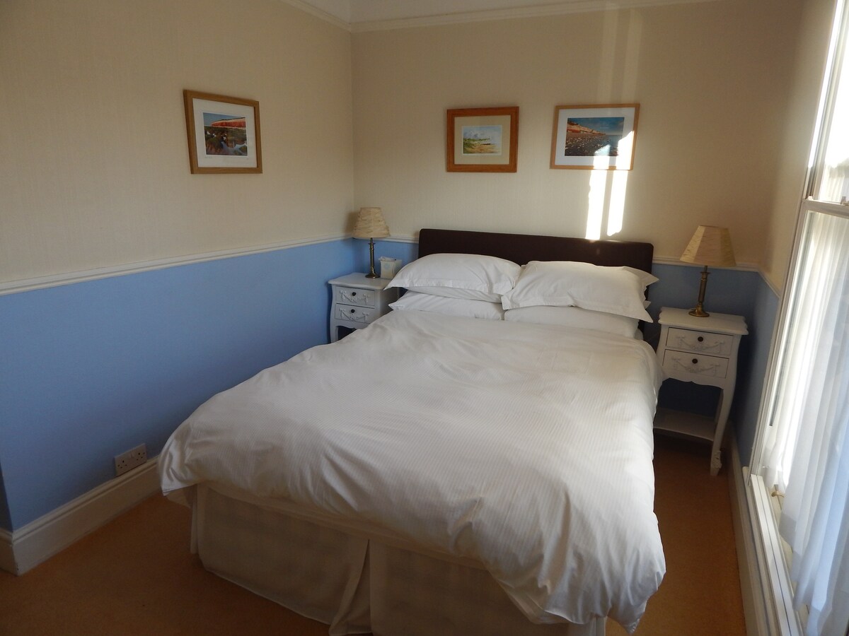 A well-lit bedroom features a spacious bed with crisp white linens. Two bedside tables with lamps are placed on either side of the bed. Two framed artworks adorn the light-colored walls, and sunlight filters through partially drawn curtains.