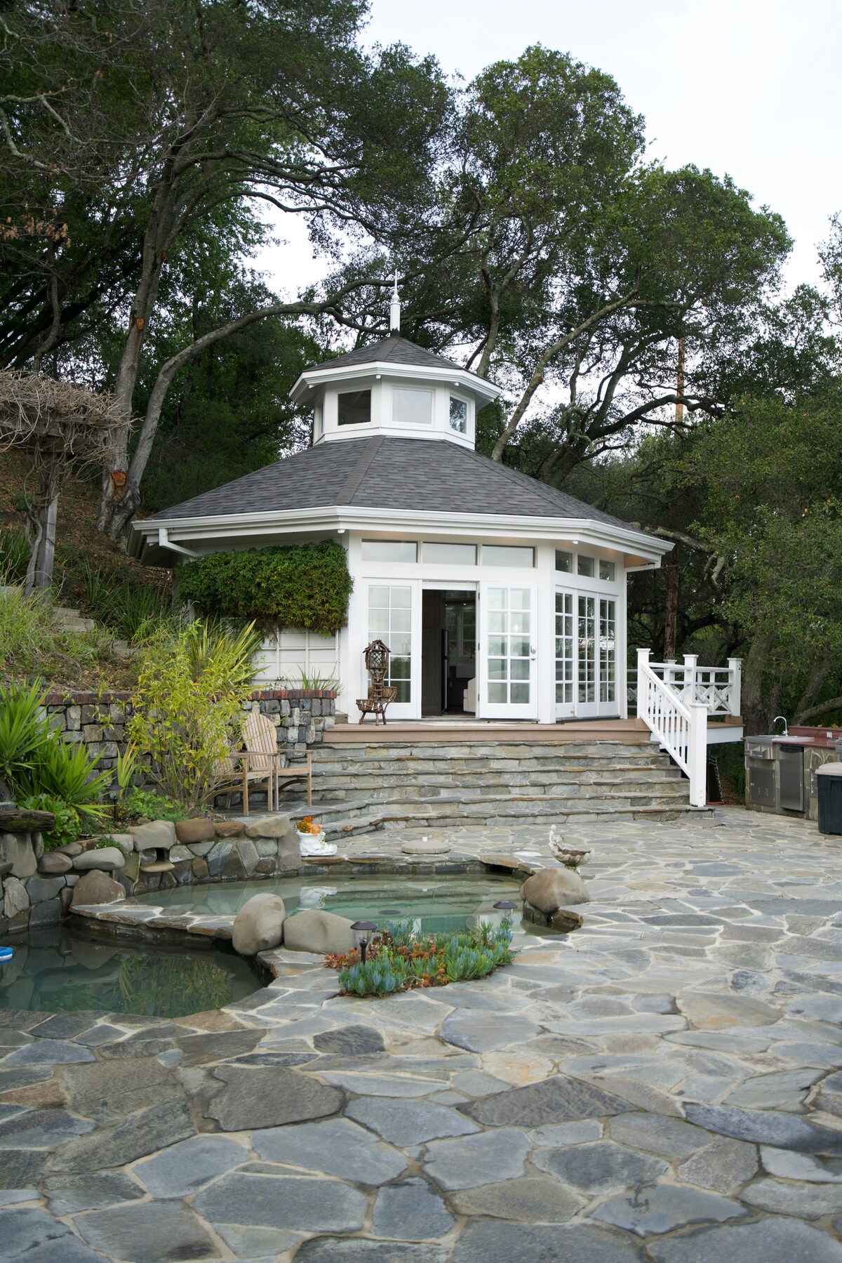 The image shows a charming cabana surrounded by greenery, featuring large windows that invite natural light. Stone steps lead up to the entrance, which is adorned with white railings. A tranquil pond with rocks is visible in the foreground, adding a serene touch to the outdoor setting.