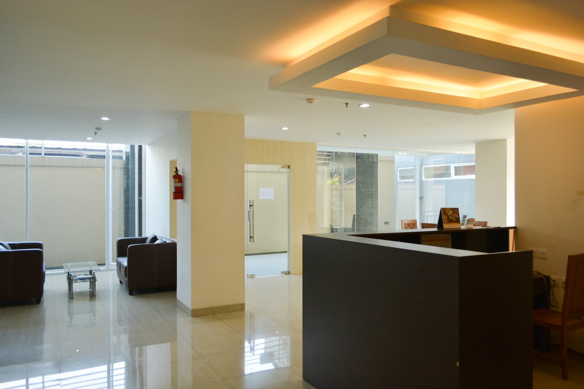 A modern lobby area features sleek tile flooring and soft lighting. Comfortable brown sofas are arranged around a glass coffee table. A reception desk is positioned to the right, while large windows allow natural light to fill the space.