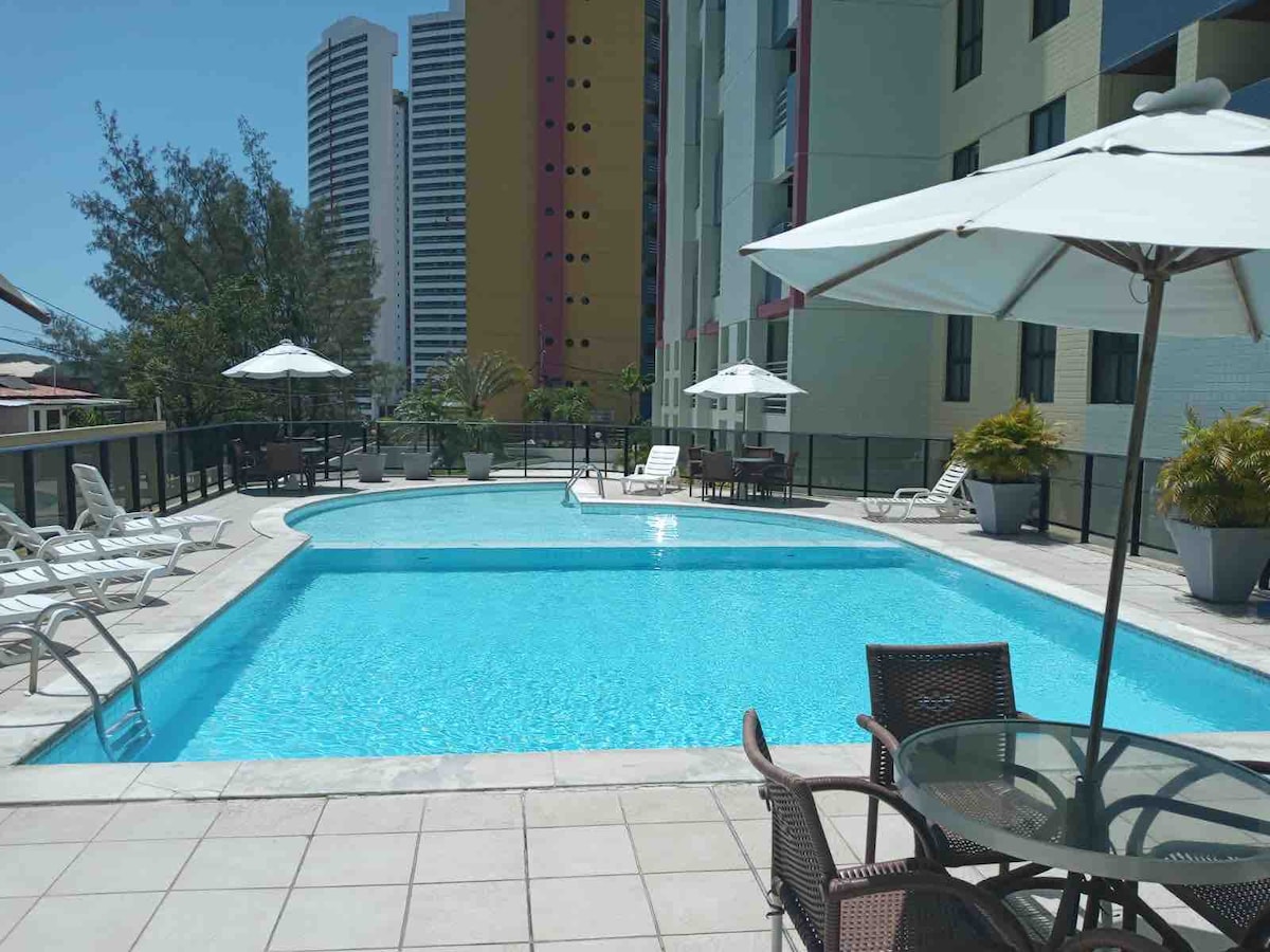 An outdoor pool area is presented, featuring a large, clear blue swimming pool surrounded by lounge chairs and patio umbrellas. Tables with chairs are arranged nearby, while tall buildings are visible in the background, framed by greenery.
