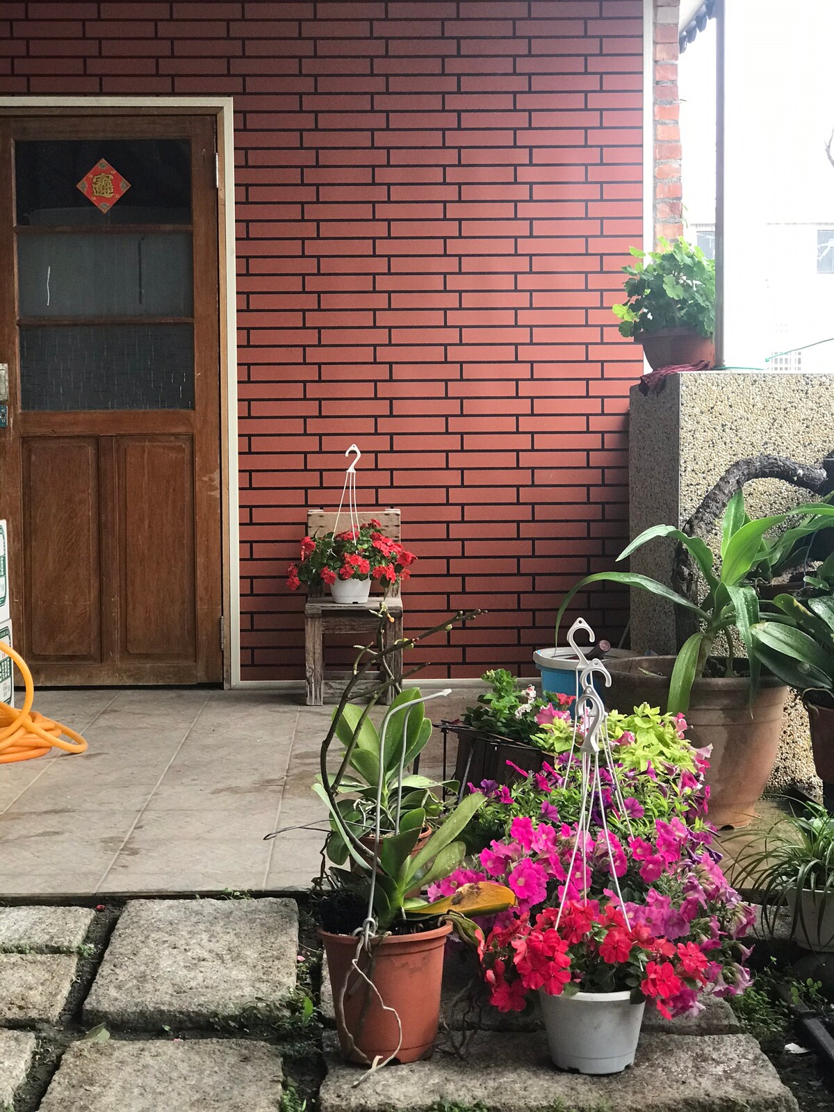 A welcoming outdoor area is presented with various potted plants, showcasing vibrant flowers in shades of pink and red. The brick wall serves as a backdrop to the entrance, while a wooden door adds a traditional touch. A stone pathway leads through the garden space.