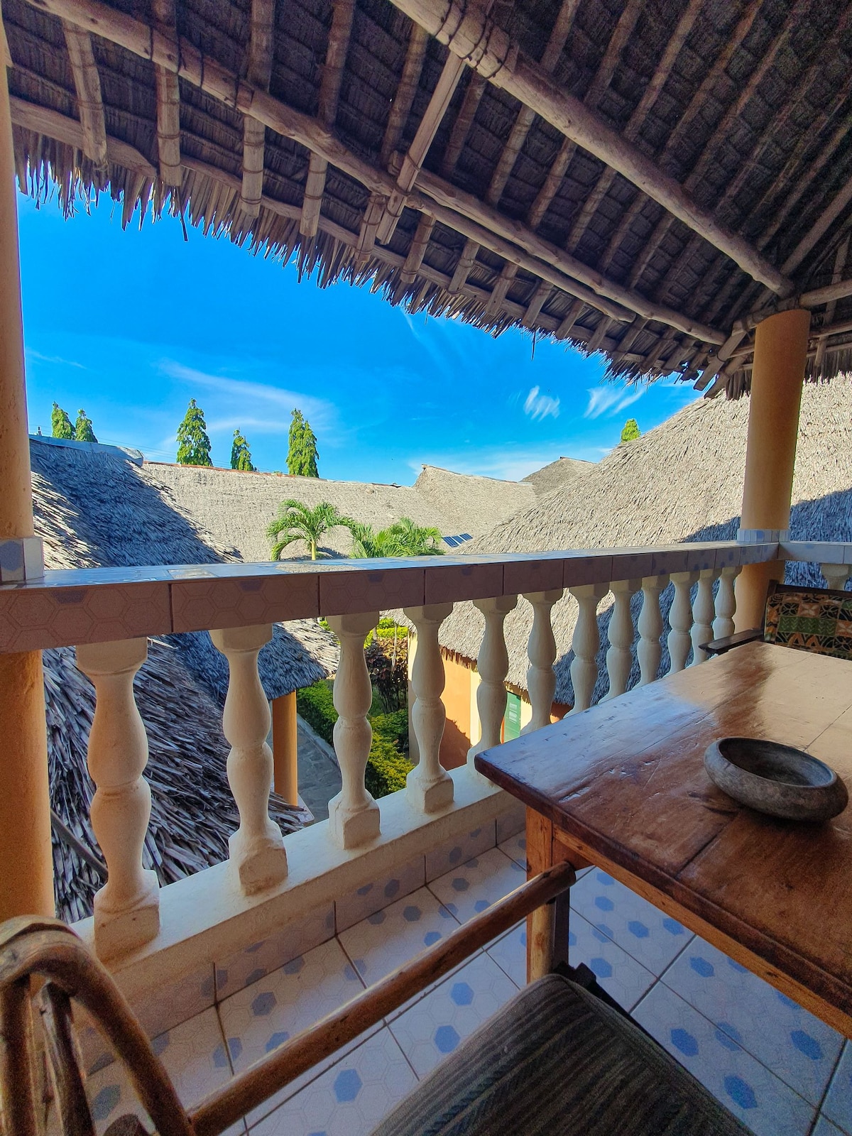 An outdoor balcony view features a low wooden table and a chair. Thatched roofs of neighboring structures and lush greenery can be seen under a bright blue sky, indicating a peaceful and sunny atmosphere.