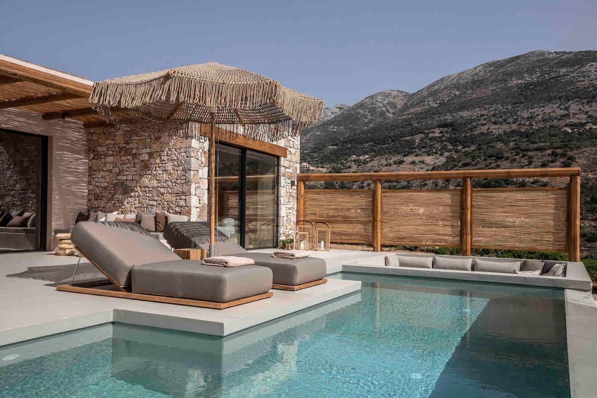 A serene outdoor space is showcased with a private swimming pool and two sun loungers positioned under a decorative canopy. The surrounding stone architecture and wooden railing provide a natural feel against the backdrop of mountains.