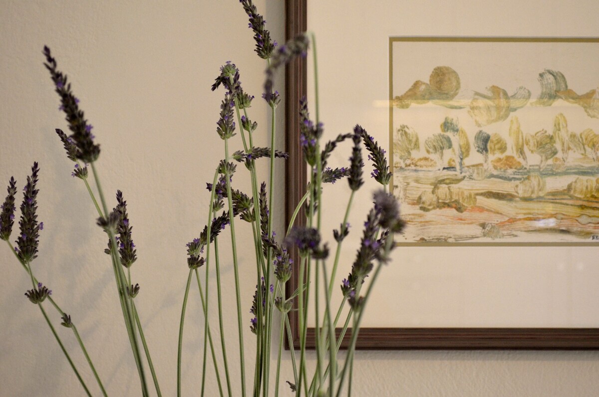 A vase with tall lavender stems is situated in front of a framed artwork, depicting a serene landscape scene. The gentle purple hues of the lavender contrast with the warm tones of the artwork and the neutral wall background.
