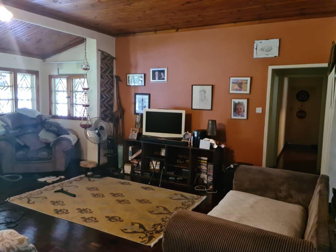 A warm living room is featured with wooden ceiling beams and an orange accent wall. A television is placed on a dark wooden cabinet, flanked by decorative items. Two couches, one beige and one brown, are arranged around a large area rug, contributing to a comfortable atmosphere.