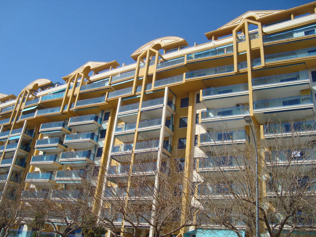 A multi-story residential building is visible against a clear blue sky. The structure features multiple balconies with glass railings, enhancing the modern architectural design. Leafless trees line the foreground, adding greenery to the scene while emphasizing the building's prominent height and form.