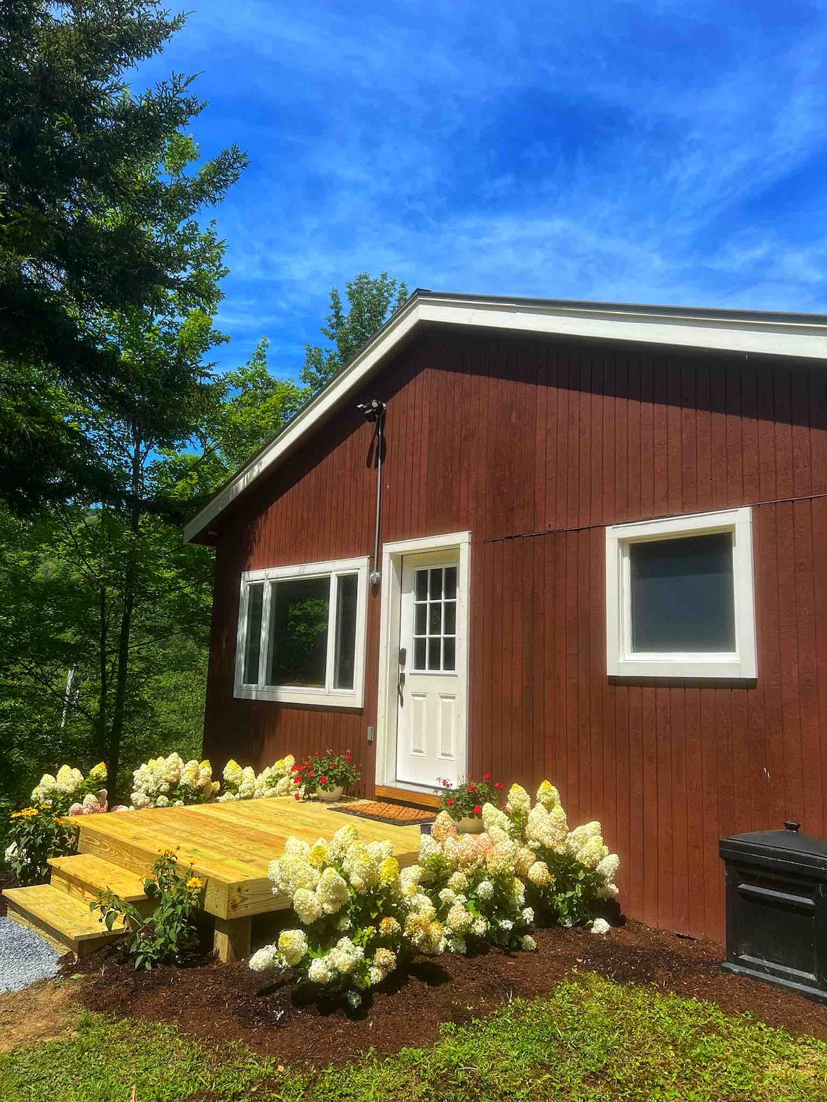 A charming red cabin is set against a backdrop of lush greenery, featuring a welcoming front porch with wooden steps. Bright white flowers bloom in neatly arranged flower beds, complementing the home's exterior. Clear blue skies enhance the tranquil atmosphere of the property.