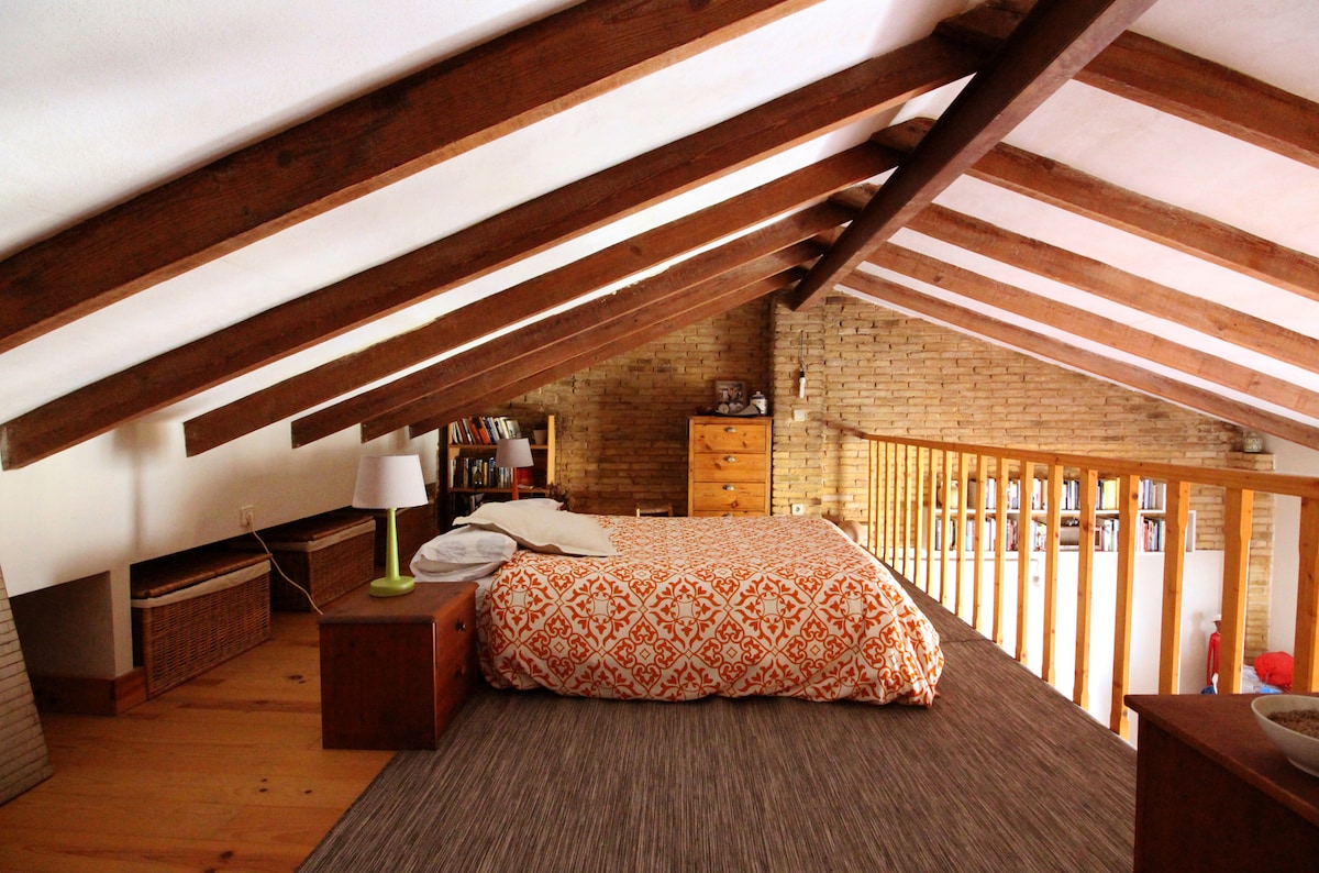 An open loft sleeping area features a spacious bed adorned with an orange and white patterned duvet. Exposed wooden beams add warmth, while natural light filters through nearby windows. A wooden railing overlooks the living space below, providing a sense of openness.