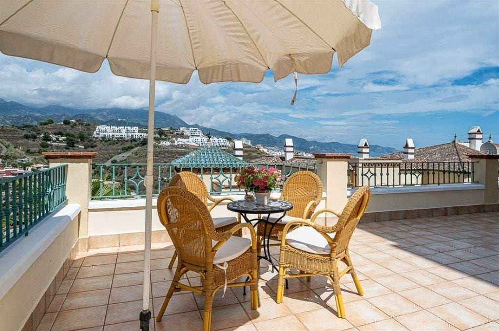A spacious terrace is shown with a round table surrounded by two wicker chairs, providing a comfortable seating area. An umbrella offers shade, and a small plant in the center adds a touch of greenery. Scenic views of mountains and rooftops are visible in the background.