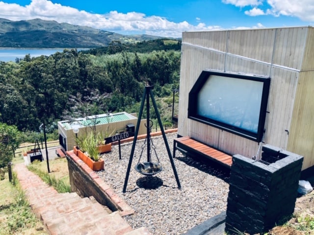 A glamping site is presented with a cozy outdoor seating area, featuring a swing seat and gravel flooring. The backdrop includes verdant hills and a shimmering lagoon in the distance. The container structure is tastefully designed with large windows, inviting natural light.