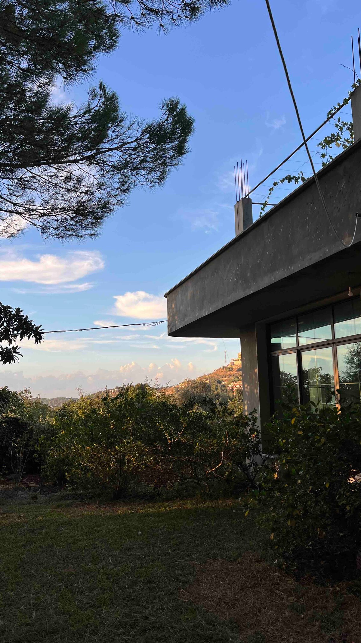 The exterior of the guesthouse is viewed from a distance, showcasing a modern architectural design with large windows. Lush greenery surrounds the property, and a clear blue sky is visible above, hinting at the natural beauty of the surrounding landscape.