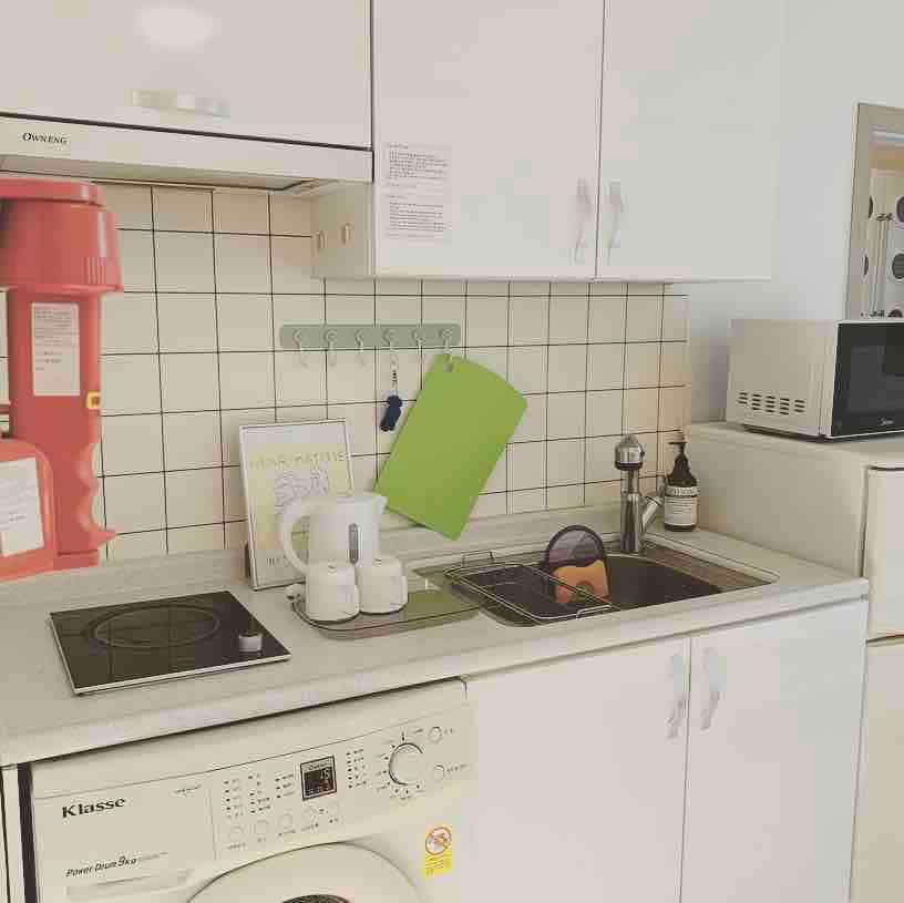 A compact kitchen area is presented, featuring a stove, sink, and washing machine. Essential kitchen items, including utensils and dishware, are neatly arranged. A microwave is positioned above the countertop, and a bright green cutting board is also visible. The environment appears well-organized and functional.