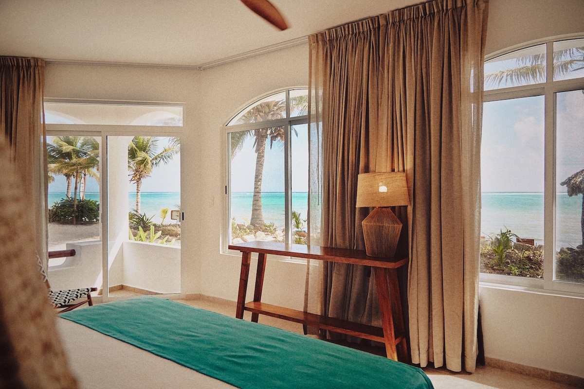 A bright room features large windows with sweeping ocean views, framed by light, flowing curtains. A wooden table stands beside the spacious bed, topped with a geometric lamp that adds a modern touch. Tropical palm trees are visible outside, enhancing the coastal ambiance.