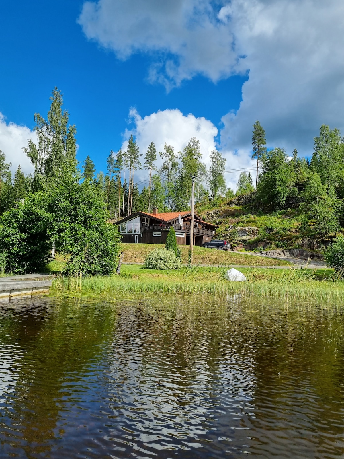 A rustic house with a red roof is nestled among lush greenery, set against a backdrop of tall trees and a bright blue sky. The tranquil water reflects the surrounding landscape, enhancing the home's serene and secluded atmosphere.