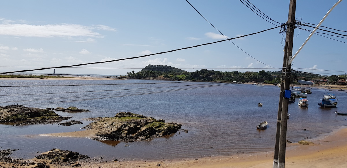 A scenic view of a river estuary is displayed, featuring rocky outcrops and a sandy shore. Small boats are anchored on the calm water, with lush greenery visible in the distance. Telephone poles and wires are positioned in the foreground, framing the landscape.