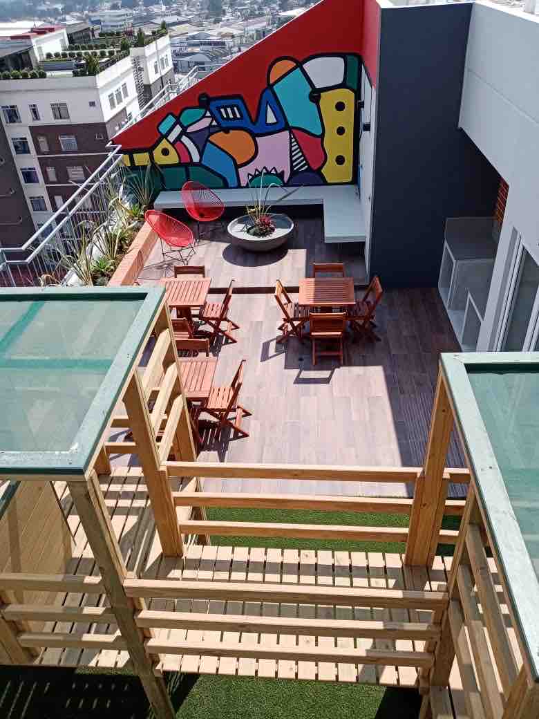 An outdoor terrace is visible, featuring a modern mural on the wall. Wooden dining tables and chairs are arranged around a circular stone table. Bright red chairs provide additional seating. Planters with greenery add a fresh touch to the urban space.