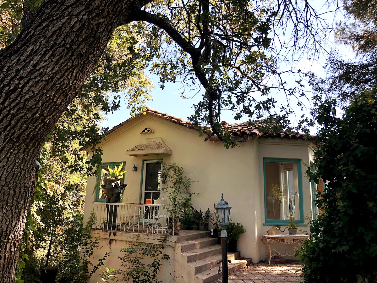 The entrance to the suite is framed by a large, leafy tree, with steps leading up to a charming patio. A decorative balcony overlooks the surrounding greenery, while potted plants add a touch of nature. The building showcases a Mediterranean design with a tiled roof and colorful window frames.