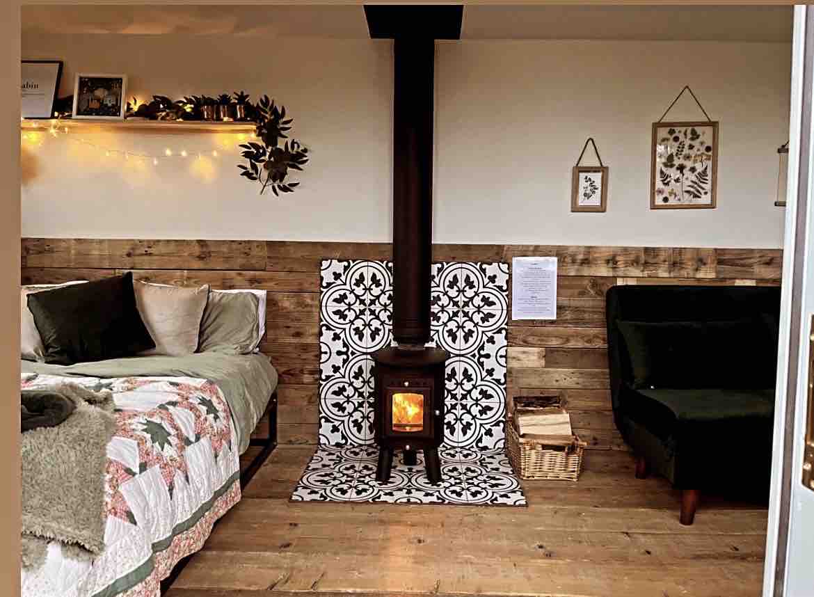 The interior features a cozy sleeping area with a quilt-covered bed and a velvet sofa, complemented by wooden walls. A wood-burning stove stands prominently on a decorative tiled base, with warm light visible through its glass door. Decorative wall art and soft lighting enhance the relaxed ambiance.