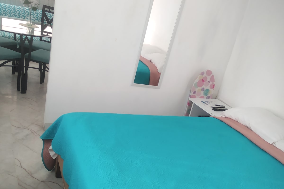 A spacious sleeping area features a vibrant blue bedspread draped over a large bed. A mirror is mounted on the wall nearby, and a small nightstand holds essentials. In the background, a dining area with a patterned table and chairs can be seen.