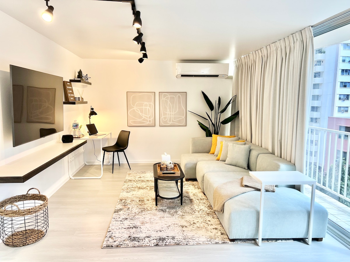 A spacious living area showcases a comfortable sofa adorned with accent pillows. A coffee table sits in front, accompanied by a neutral area rug. A desk with task lighting and a stylish chair provide a work space. Large windows are framed by light curtains, enhancing natural light.