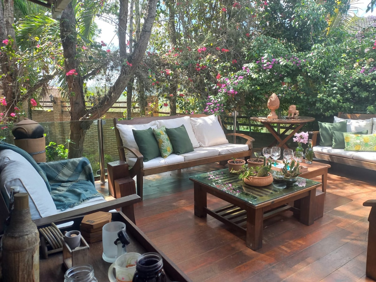 An outdoor seating area features wooden furniture arranged around a coffee table, surrounded by lush greenery and flowering plants. Cushions in soft tones add comfort, and a variety of decorative elements, including potted plants and dishes, enhance the inviting atmosphere.