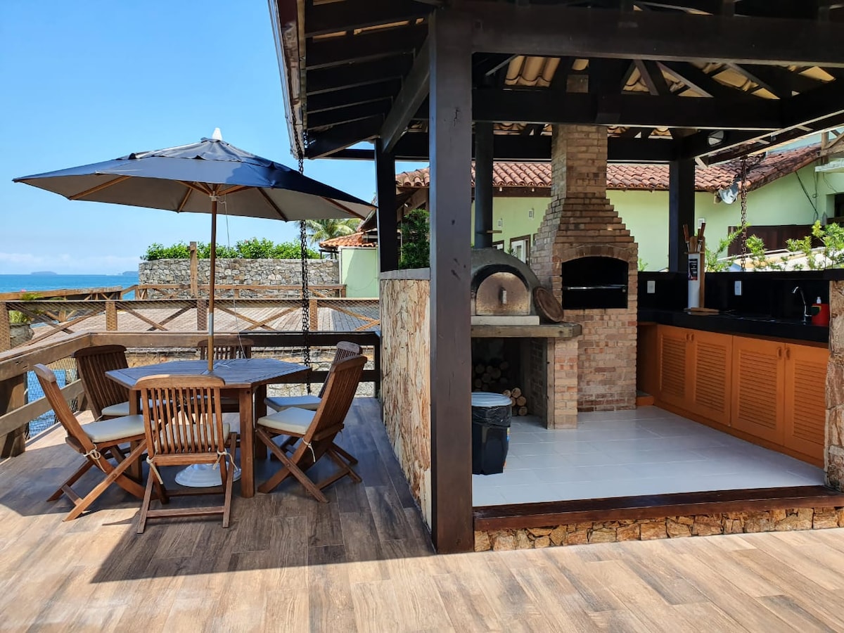A covered outdoor kitchen features a brick oven and ample counter space, complemented by wooden cabinetry. A dining area with a round table and four chairs is shaded by a patio umbrella. Scenic views of the sea and surrounding landscape can be observed in the background.