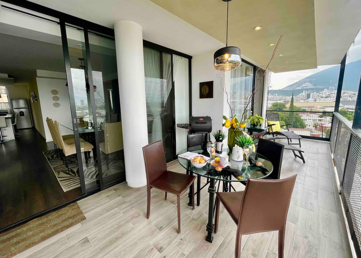A glass dining table is set for four, surrounded by dark chairs with modern design. Potted plants and fresh flowers adorn the table, with a scenic view of the city visible through the large sliding glass doors that open to the living area.
