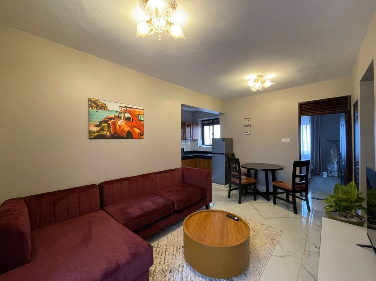 A cozy living area features a maroon sectional sofa and a wooden coffee table. A round dining table with four chairs is positioned nearby. A painting depicting a vintage car adds a pop of color to the soft-toned walls, while natural light filters through the window.