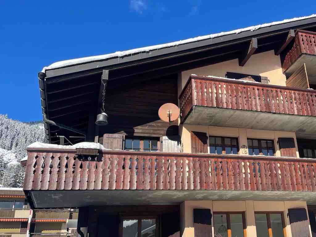 The exterior of the chalet is showcased, featuring wooden balconies with decorative railings. A satellite dish is mounted on the side, and the sloped roof is partially covered in snow. The backdrop includes trees blanketed in snow, under a clear blue sky.
