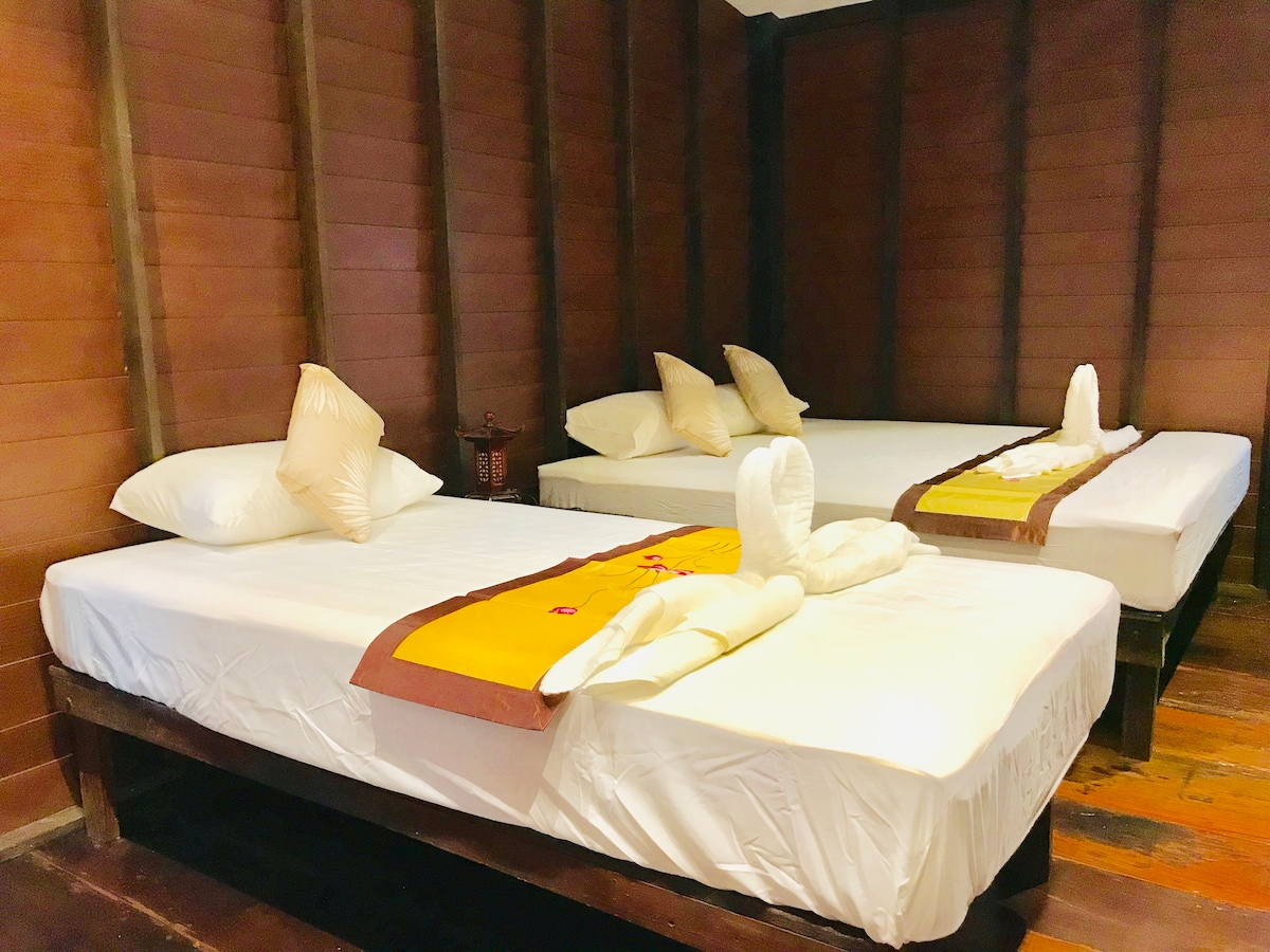 A cozy sleeping area features two spacious beds, each adorned with soft pillows. Simple decorative elements include folded towels arranged artistically. Rich wooden walls contribute to a warm ambiance, and natural light filters in, enhancing the room's welcoming feel.